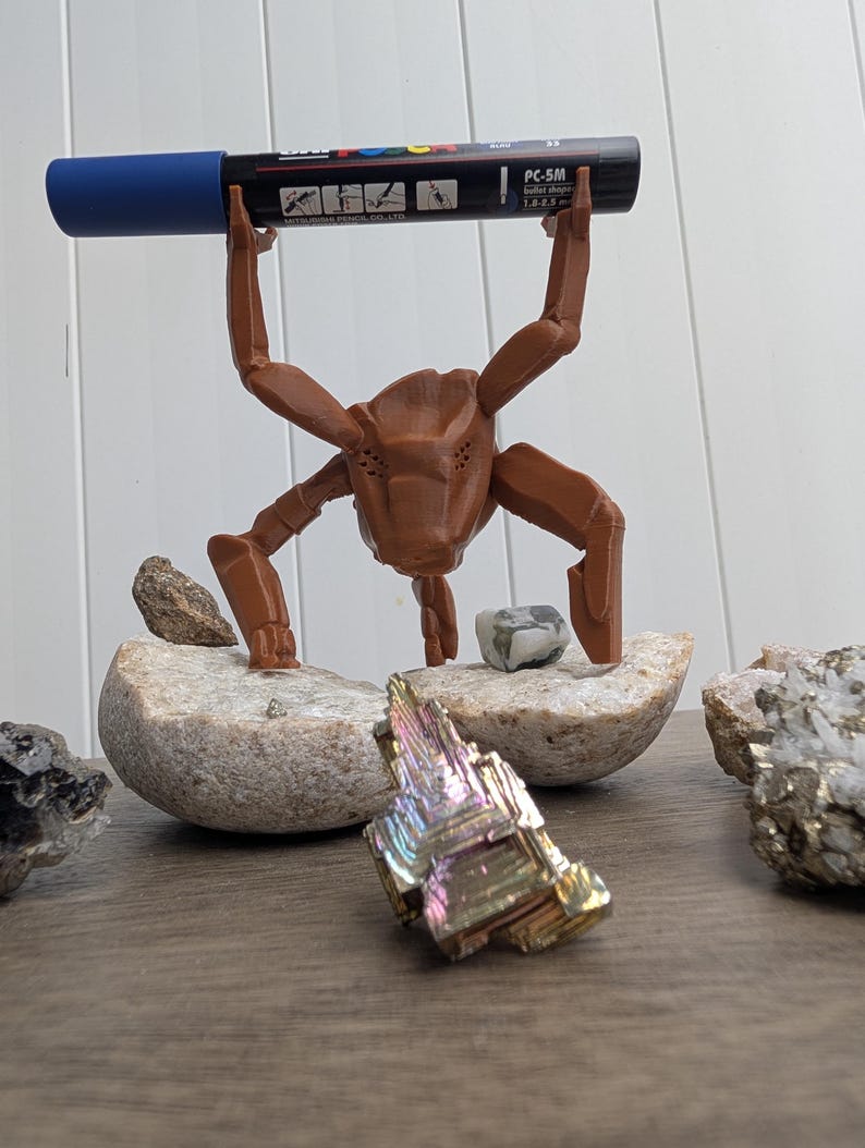 May include: A brown 3D-printed insect-like sculpture holding a blue and black marker labeled "PC-5M". The sculpture is positioned on a stone, with colorful crystals and other stones nearby. The background is a light-colored wall.