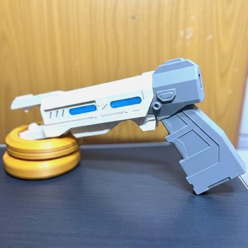 May include: A futuristic toy blaster featuring a white barrel with blue rectangular accents and a gray handle. The blaster is supported by a circular, golden-yellow base. The design includes geometric paneling and a blocky, angular construction.