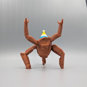 May include: A brown, 3D-printed creature with six legs and arms raised in the air. It wears a party hat with blue, yellow, and white accents. The creature has a textured surface and is standing on a gray surface.