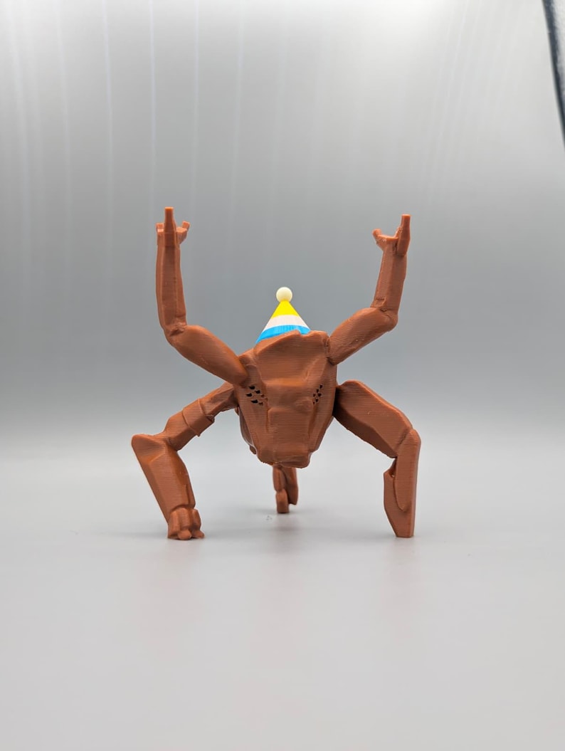 May include: A brown, 3D-printed creature with six legs and arms raised in the air. It wears a party hat with blue, yellow, and white accents. The creature has a textured surface and is standing on a gray surface.