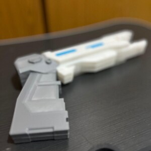 May include: A toy blaster featuring a gray handle and a white barrel section. The barrel includes a rectangular blue accent detail. The object has a geometric, angular design with a prominent grip and a trigger guard area visible in the foreground.