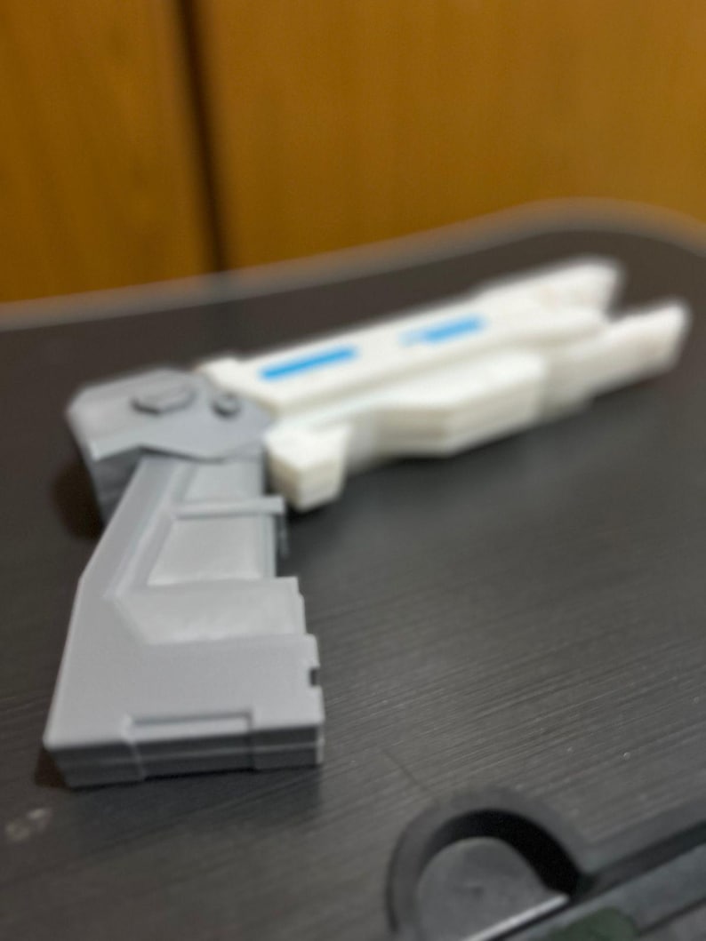May include: A toy blaster featuring a gray handle and a white barrel section. The barrel includes a rectangular blue accent detail. The object has a geometric, angular design with a prominent grip and a trigger guard area visible in the foreground.