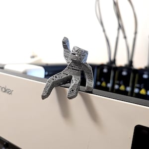May include: A small, gray, 3D-printed figurine with a blocky, humanoid shape and a complex, open-lattice geometric texture. The figure is posed in a seated position with one arm raised, featuring a hollow, intricate structure throughout its body.