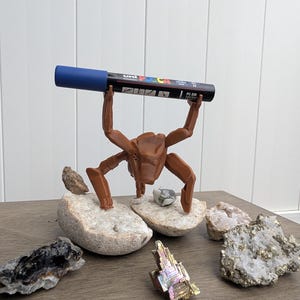 May include: A brown, crab-shaped pen holder supporting a black and blue Uni Posca PC-5M marker. The pen holder is on a wooden surface, surrounded by various rocks and minerals, including quartz and pyrite.