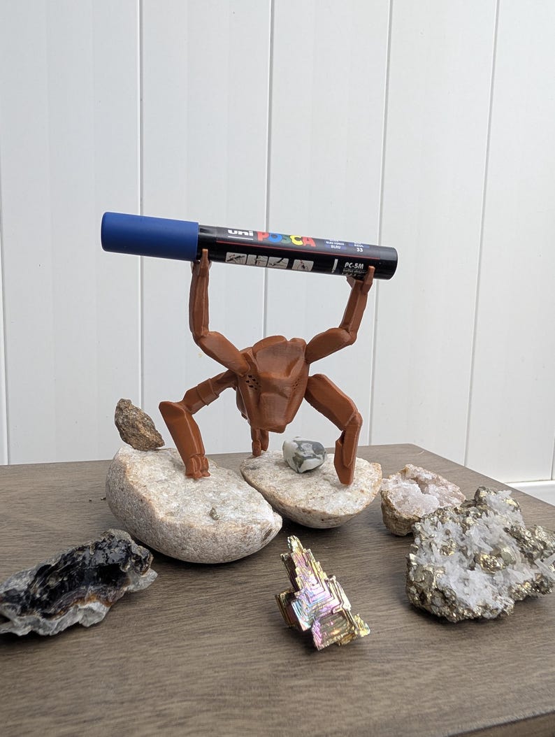 May include: A brown, crab-shaped pen holder supporting a black and blue Uni Posca PC-5M marker. The pen holder is on a wooden surface, surrounded by various rocks and minerals, including quartz and pyrite.