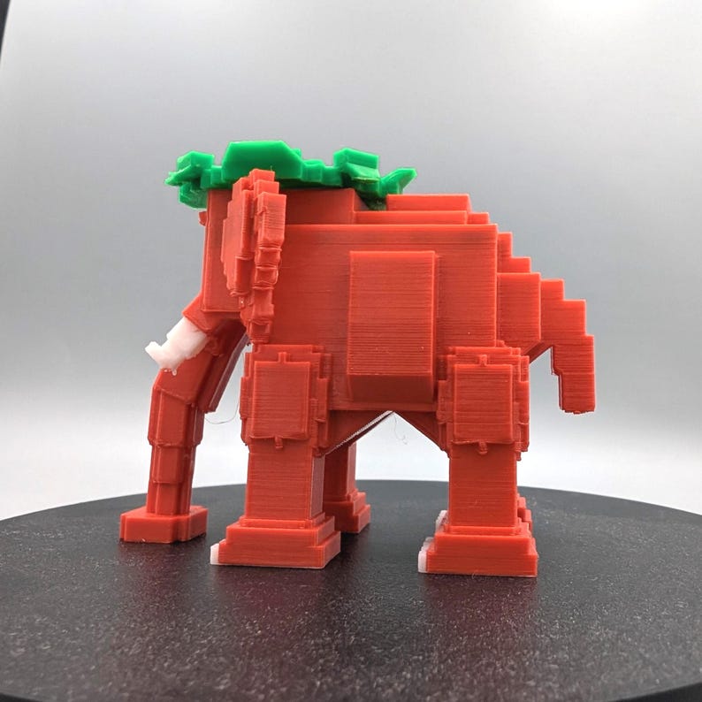 May include: A 3D-printed, pixelated elephant figurine in shades of red, with green accents on its head and white tusks and feet. The geometric design gives it a retro, video game aesthetic. The elephant is standing on a black surface.