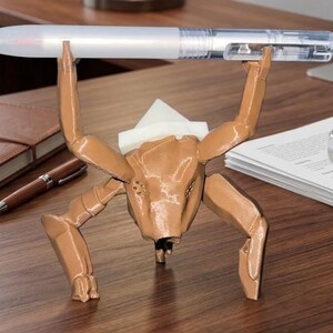 May include: A brown, insect-like pen holder with a pen held in its arms. The pen holder is made of a 3D-printed material and has a unique, artistic design. The pen is white and clear.