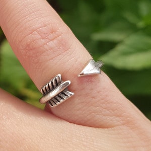 Könnte beinhalten: Ein silberner Pfeilring mit einem detaillierten Design. Der Ring ist offen und verstellbar, um verschiedenen Fingergrößen zu passen.