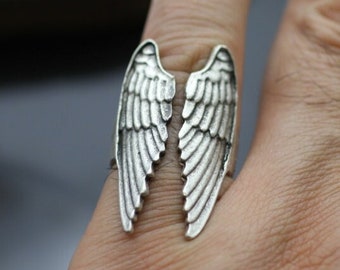 May include: A silver-colored ring featuring a detailed design of two open wings. The wings are positioned to appear as if they are wrapping around the finger. The ring is worn on a hand, showcasing the intricate feather detailing.