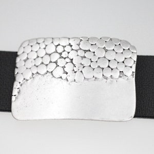 May include: A silver belt buckle with a textured surface of small circles. The buckle is attached to a black leather belt.