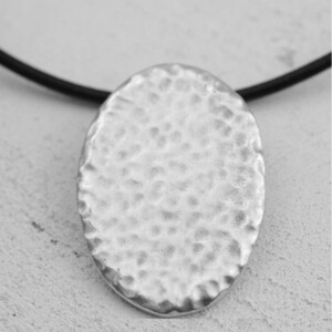 May include: A silver oval pendant necklace with a hammered texture. The pendant is attached to a black cord.