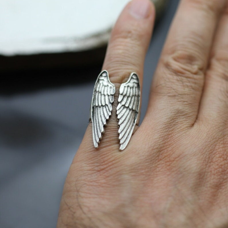 May include: A silver-colored ring featuring a detailed design of two open wings. The wings are positioned to appear as if they are wrapping around the finger. The ring is worn on a hand, showcasing the intricate feather detailing.