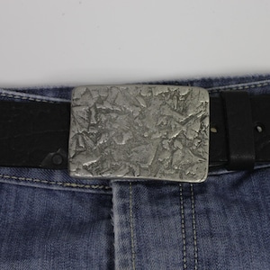 May include: A silver belt buckle with a textured, abstract design. The buckle is attached to a black leather belt and is worn on a pair of blue jeans.