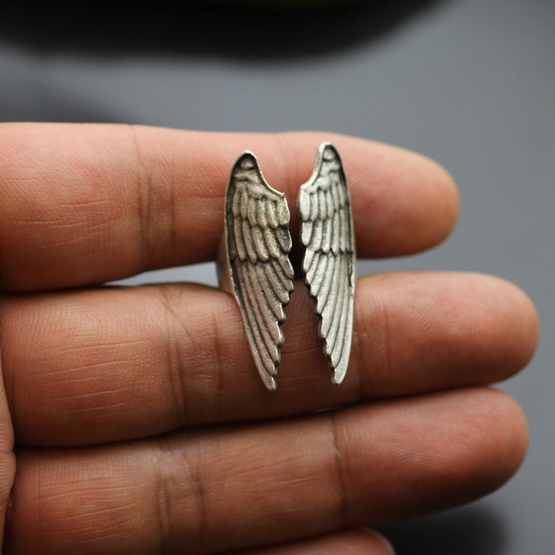 May include: A silver-toned ring featuring a split wing design. The ring's wings are detailed with feather textures, creating a three-dimensional effect. The ring is designed to be worn on a finger.