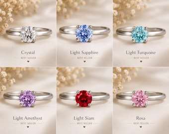 May include: A collection of six silver-toned rings, each featuring a single round-cut gemstone in different colors: clear crystal, light sapphire blue, light turquoise, light amethyst purple, light siam red, and pink. Each ring is labeled below.