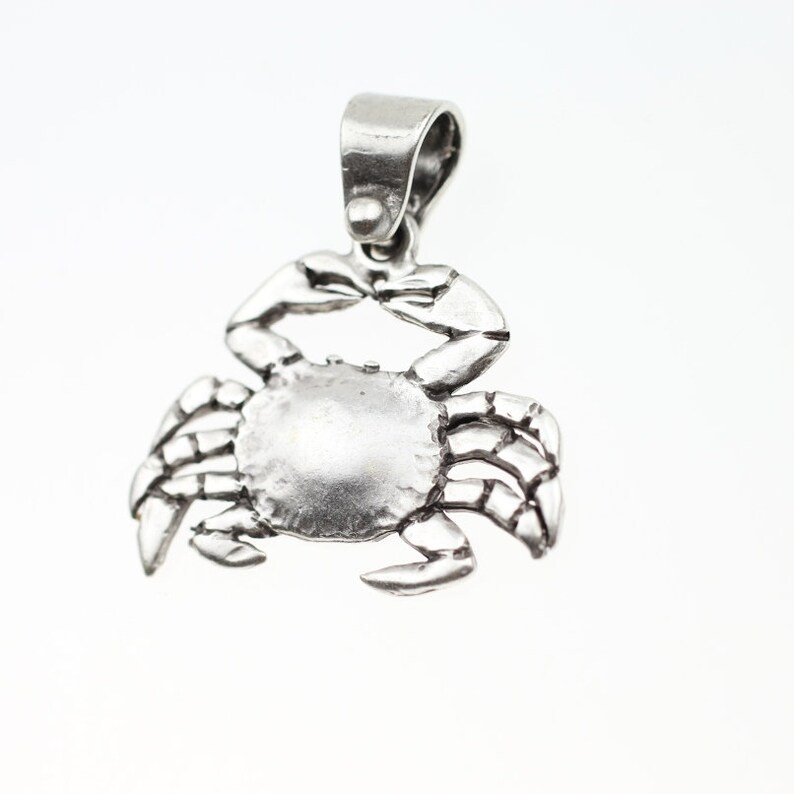 May include: A silver crab charm pendant. The crab is detailed with textured legs and a smooth, rounded body.