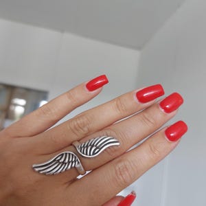 K&ouml;nnte beinhalten: Ein silberfarbener Ring mit einem Fl&uuml;geldesign. Der Ring hat zwei fl&uuml;gelf&ouml;rmige Elemente, eines auf jeder Seite des Fingers. Der Ring wird an einer Hand mit rotem Nagellack getragen.