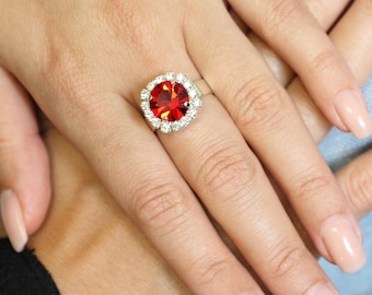 May include: A silver ring featuring a large, round, red gemstone surrounded by smaller, clear stones. The ring is set on a hand with manicured nails, showcasing the jewelry's design and color contrast.