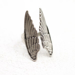 May include: A silver-toned ring featuring a detailed wing design. The ring has two open wings, each with intricate feather detailing. The ring is set against a white background, highlighting the metallic finish and the ring's unique shape.