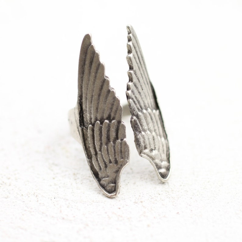 May include: A silver-toned ring featuring a detailed wing design. The ring has two open wings, each with intricate feather detailing. The ring is set against a white background, highlighting the metallic finish and the ring's unique shape.
