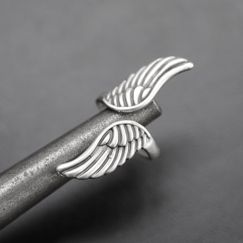 K&ouml;nnte beinhalten: Ein silberfarbener Ring mit detaillierten Fl&uuml;gelmotiven. Die Fl&uuml;gel sind auf gegen&uuml;berliegenden Seiten des Rings angeordnet. Der Ring wird auf einem grauen Metallstab vor grauem Hintergrund pr&auml;sentiert. Die Fl&uuml;gel sind detailliert.
