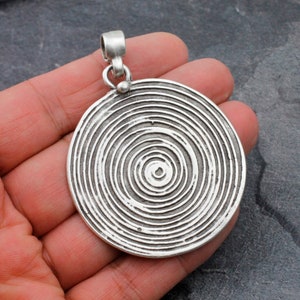 May include: Silver pendant with a circular design featuring concentric circles. The pendant has a loop at the top for attaching to a chain.