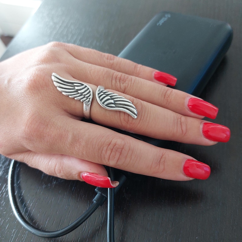 K&ouml;nnte beinhalten: Ein silberner Ring mit einem Fl&uuml;geldesign. Der Ring hat zwei detaillierte Fl&uuml;gel, einen auf jeder Seite des Fingers. Der Ring wird an einer Hand mit rotem Nagellack getragen.