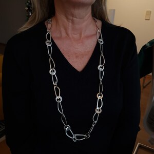 Puede incluir: Un collar largo plateado con una mezcla de eslabones ovalados y circulares. El collar se lleva sobre una camiseta negra con cuello en V. Los eslabones alternan entre óvalos alargados y anillos circulares, creando un diseño geométrico moderno.