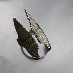 May include: A silver-toned ring with a wing design. The ring features two detailed wings that extend outwards, creating an open design. The wings have a textured surface with feather-like detailing. The ring is set against a dark background.