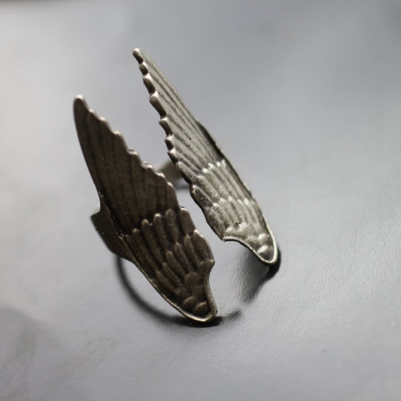 May include: A silver-toned ring with a wing design. The ring features two detailed wings that extend outwards, creating an open design. The wings have a textured surface with feather-like detailing. The ring is set against a dark background.