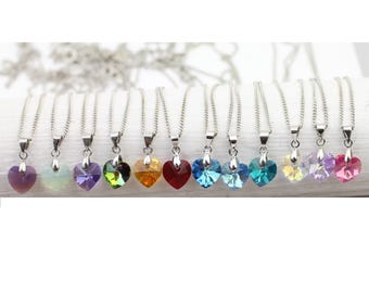 Puede incluir: Una colección de collares con colgantes en forma de corazón de varios colores, incluyendo morado, verde, naranja, rojo y azul. Cada colgante está suspendido de una cadena plateada. Los collares se exhiben sobre una superficie blanca.