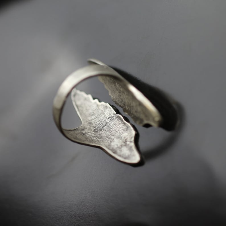May include: A silver-toned ring with an open design. The ring features two stylized, leaf-like shapes that curve around the finger. The surface of the ring has a textured appearance, and the overall design is modern and organic.