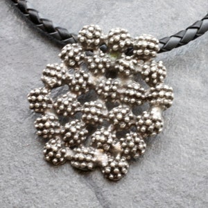 Puede incluir: Un collar con un colgante de plata con un diseño detallado e intrincado. El colgante está hecho de muchas pequeñas cuentas redondas que se agrupan para formar un patrón único. El collar está en un cordón de cuero negro.
