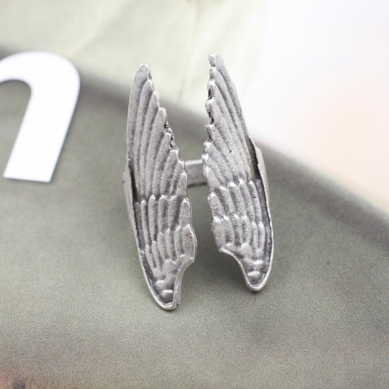 May include: A silver-toned ring featuring a detailed wing design. The ring has two separate wing shapes that extend upwards, with intricate feather detailing. The ring is likely made of metal and has a vintage or antique appearance.