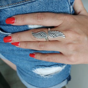 K&ouml;nnte beinhalten: Ein silberfarbener Ring mit einem Fl&uuml;geldesign. Der Ring wird an einem Finger getragen, wobei sich die Fl&uuml;gel nach au&szlig;en erstrecken. Das Design des Rings ist detailliert, wobei jede Feder deutlich sichtbar ist. Der Hintergrund umfasst Jeansshorts und eine Hand mit rotem Nagellack.