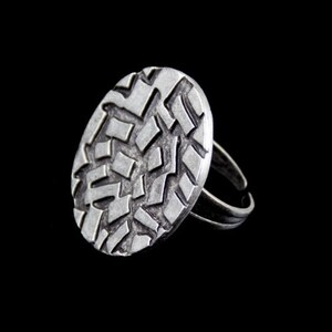 Puede incluir: Anillo plateado con una cara ovalada texturizada. La cara está cubierta con un patrón geométrico de formas rectangulares. El anillo tiene una banda ajustable. La imagen está sobre un fondo negro.