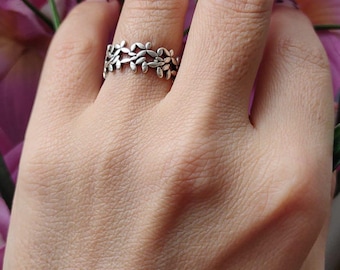 May include: A silver ring featuring a repeating leaf design. The ring is worn on a finger, with pink flowers in the background. The ring's intricate details are visible, showcasing a nature-inspired aesthetic.