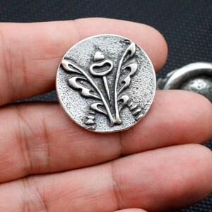 Puede incluir: Un botón ovalado de plata con un diseño en relieve de una flor estilizada con tres hojas.