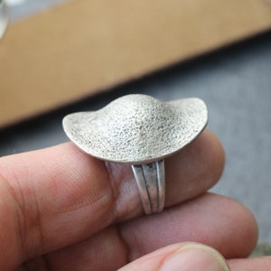 Puede incluir: Un anillo de plata con una parte superior en forma de cúpula texturizada. El anillo se lleva en un dedo.