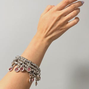 May include: Silver bracelet featuring multiple strands and dangling pink gemstones. The bracelet is worn on a wrist, highlighting its design. The bracelet has a vintage or bohemian style, with a detailed design.