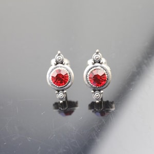 May include: A pair of silver earrings with red crystal stones. The earrings have a delicate design with small circles and a pointed top.