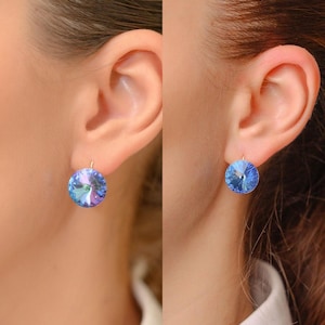 Op de afbeelding: Een paar ronde, blauwe kristallen oorbellen met goudkleurige haken. De oorbellen hebben een facetontwerp dat licht reflecteert en een sprankelend effect creëert. De oorbellen worden getoond op het oor van een persoon.