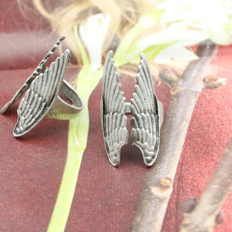 May include: Two silver-toned rings with a wing design. One ring features a single wing, while the other has two wings. The rings have a vintage, antique appearance. The wings are detailed with feather-like textures.