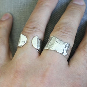 Op de afbeelding: Twee zilveren ringen met een textuurrijk, organisch ontwerp. Eén ring is open en wikkelt zich om de vinger, terwijl de andere een massieve band is met een ruw, ongelijkmatig oppervlak. De ringen worden op een hand getoond.