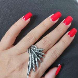 May include: A silver ring with a detailed design resembling a stylized plant or flower. The ring is worn on a hand with red nail polish.