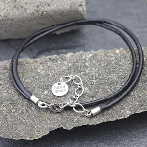 Könnte beinhalten: Eine schwarze Lederkordelkette mit einem silbernen Verschluss und einem kleinen silbernen Anhänger mit der Aufschrift "hand made".