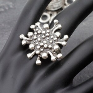 May include: A silver ring with a large, abstract design. The ring is made of many small, round beads that are clustered together in a circular shape. The beads are connected by thin, silver wires that extend outwards from the center of the ring.