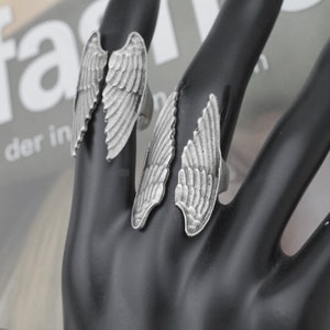 May include: Three silver-toned rings with wing designs are displayed on a black surface. Each ring features a detailed feather pattern, with the wings extending upwards. The rings are likely made of metal and are designed to be worn on fingers.