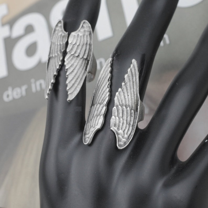 May include: Three silver-toned rings with wing designs are displayed on a black surface. Each ring features a detailed feather pattern, with the wings extending upwards. The rings are likely made of metal and are designed to be worn on fingers.