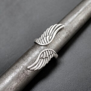 K&ouml;nnte beinhalten: Ein silberfarbener Ring mit offenem Design, der zwei stilisierte Fl&uuml;gelmotive aufweist. Die Fl&uuml;gel sind mit federartigen Linien detailliert, wodurch ein strukturiertes Aussehen entsteht. Der Ring wird auf einem Metallstab gezeigt.